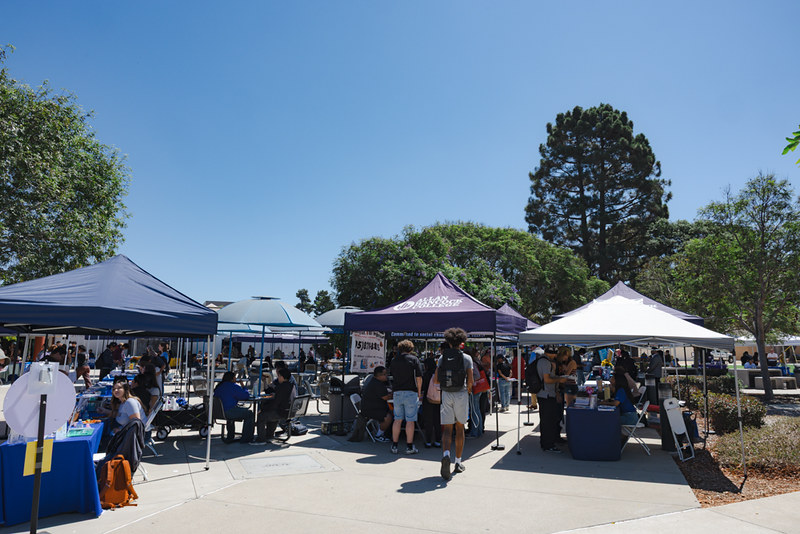 Outdoor student event at Allan Hancock College on a sunny day, with several canopy tents set up in a courtyard area. Students are gathered at tables under the tents, talking with representatives, collecting information, and enjoying the event. Large trees and clear blue sky are visible in the background, creating a lively and welcoming campus atmosphere.